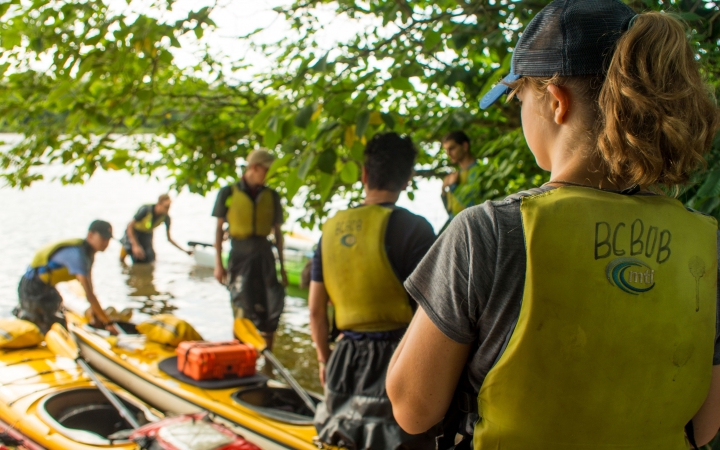 a group of young people wearing life jackets stand near kayaks beached on the shore 